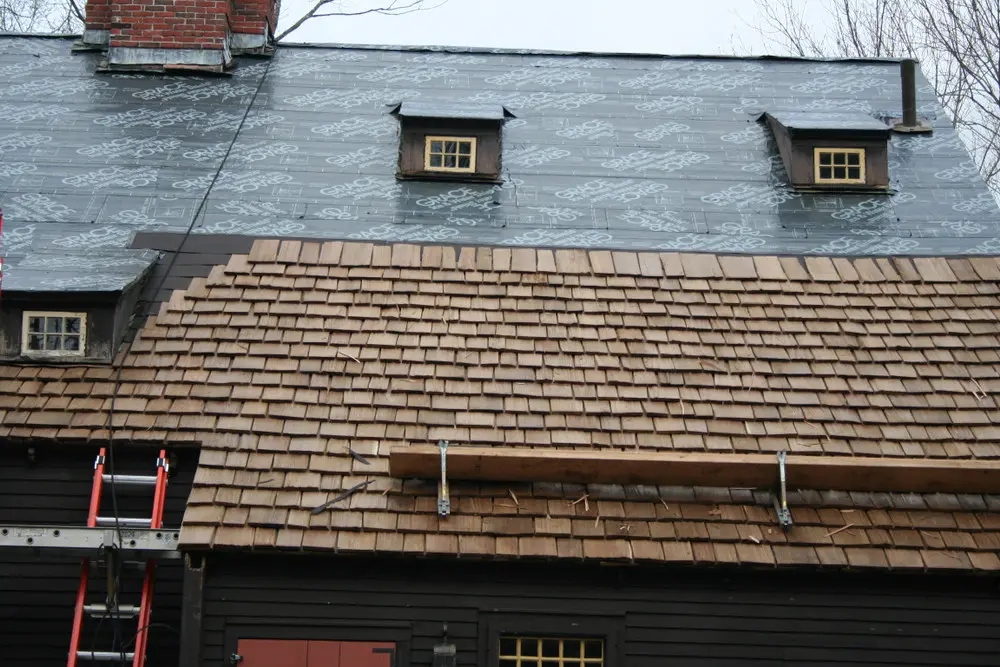 Partially installed cedar shake roof with exposed underlayment, small dormers, and a staging platform.