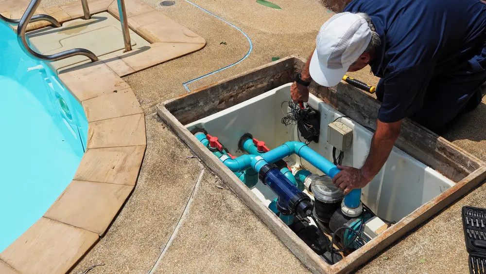 Pool technician servicing a swimming pool pump beside an in-ground pool.